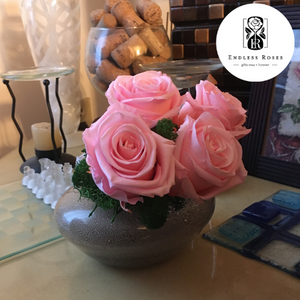 Frontal Profile. 4 extra large pink preserved roses nestled on green preserved moss displayed on pearl-shape copper glass vase on a side table in front of a chessboard and a wine-glass full of decorative corks. 