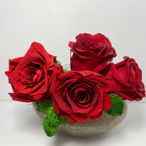 Frontal. 4 extra large red preserved roses nestled on green preserved moss displayed on pearl-shape copper glass vase